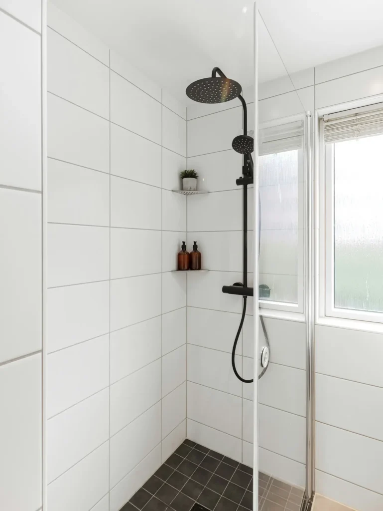 White tile shower in Burlington with black rainfall showerhead, black thermostatic faucet, glass shower panel, corner shelf, and frosted window