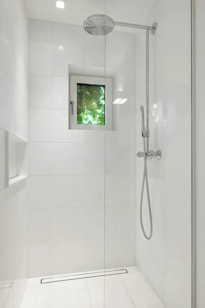 Open white tile walk-in shower in Burlington with frameless glass panel, rainfall showerhead, linear floor drain, and window