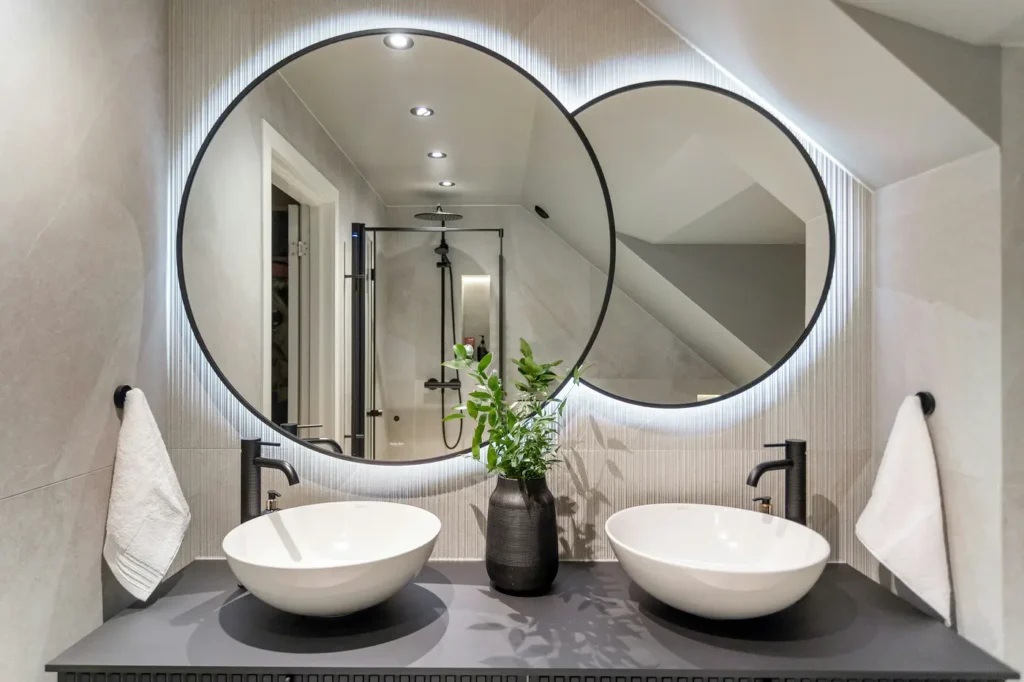 Custom bathroom design Aldershot — double vanity with backlit round mirrors, vessel sinks, and dark stone countertop