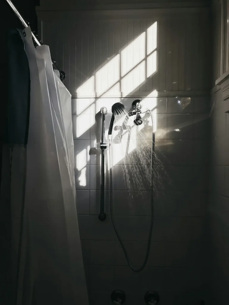 Older shower stall in Burlington with vinyl curtain, chrome handheld showerhead on slide bar, and small white tile walls