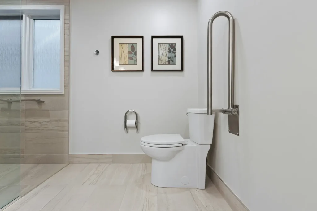 Accessible Burlington bathroom with a stainless flip-up grab bar beside the toilet, integrated toilet paper holder grab bar, and a glass shower with grab bar visible in the background