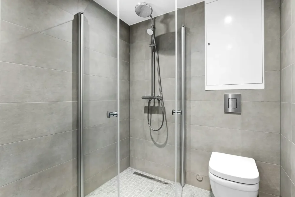 Grey large-format tile shower in Burlington with bi-fold glass door, rainfall showerhead, linear floor drain, and wall-mounted toilet