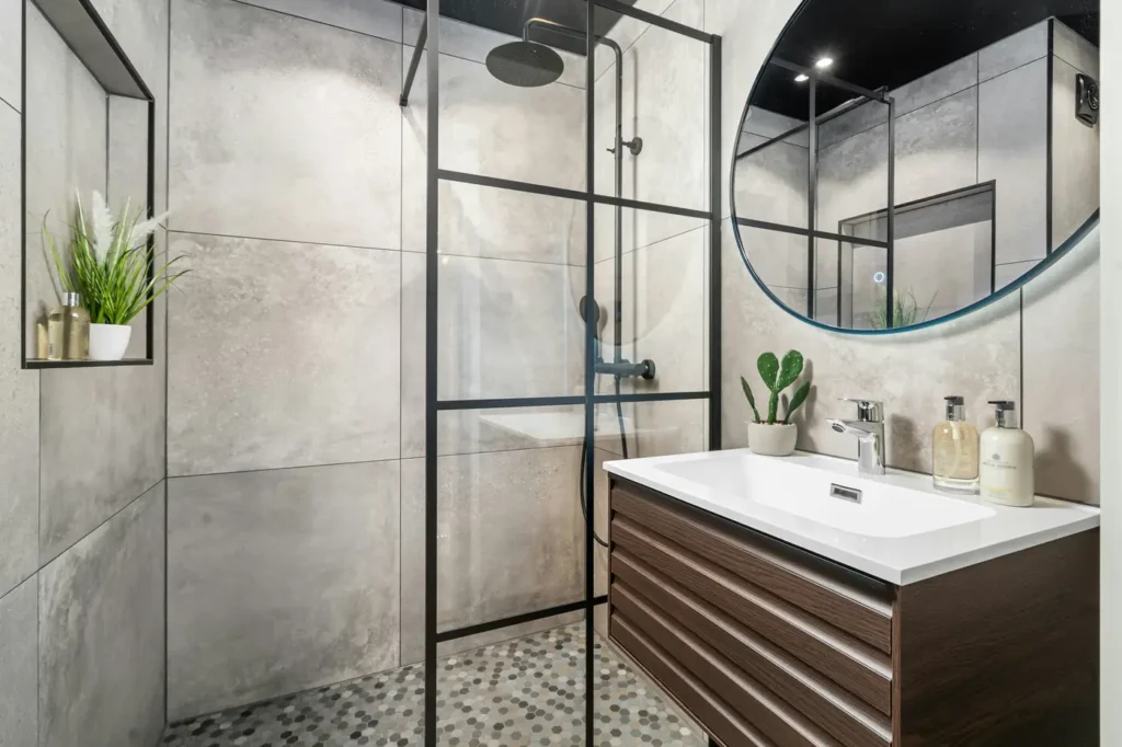 Custom walk-in shower with concrete-look tile, black-framed glass, recessed niche, and a round backlit mirror over a dark wood vanity